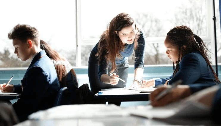 A teacher leans over a student’s desk to offer guidance during a classroom lesson. Other students in school uniforms are working at their desks in a bright, sunlit classroom.