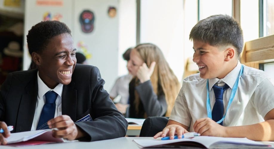 Two students in school uniforms sit at a classroom desk laughing together while working in their notebooks, with other students visible in the background.